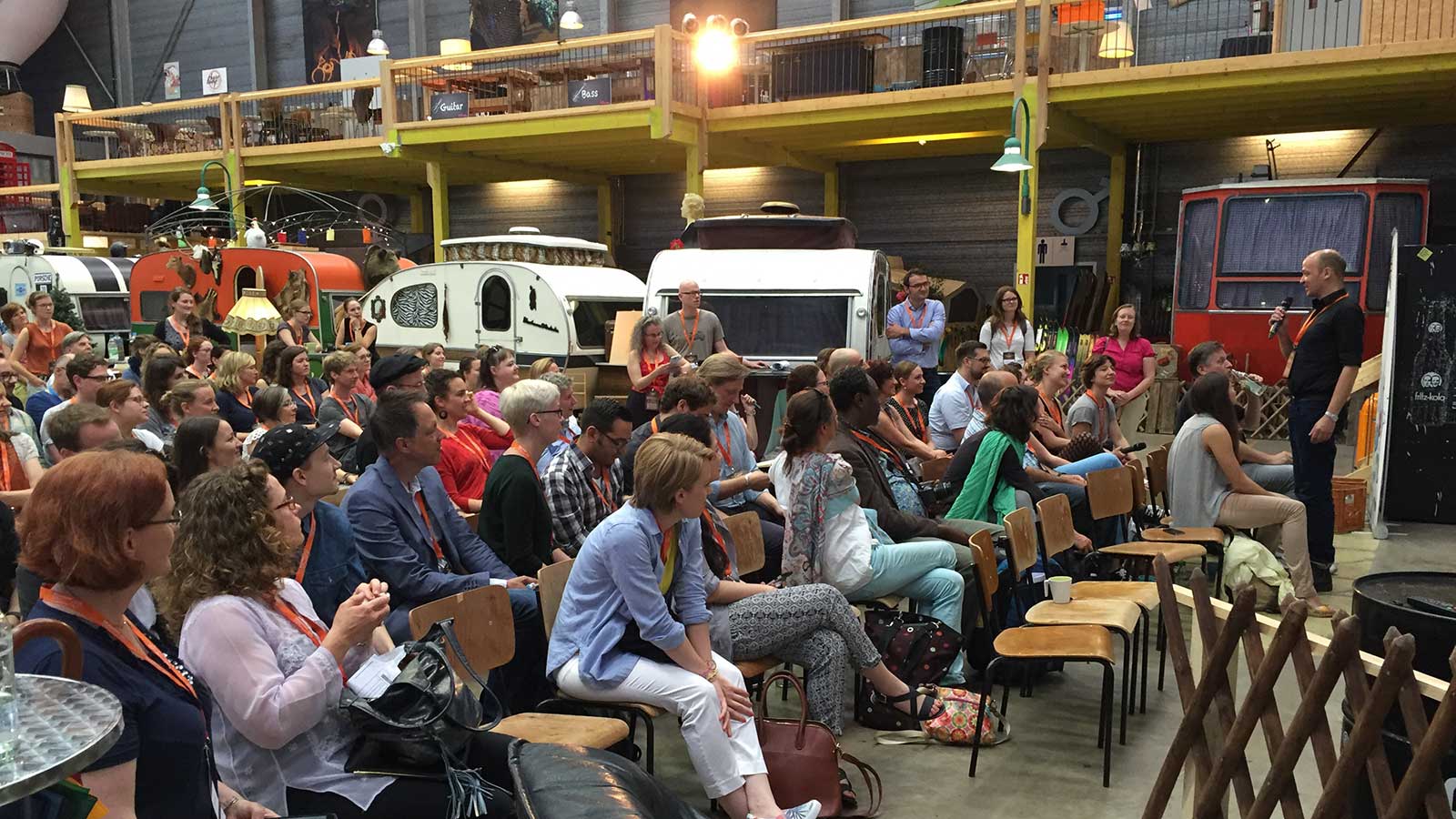 Eine größere Gruppe Menschen sitzt und hört einem Mann zu, der in ein Mikrofon spricht