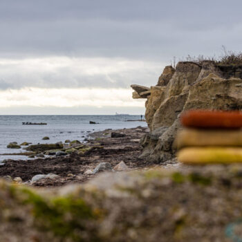 Im Vordergrund sieht man einen Steinhaufen, wo die Steine auf einander gestapelt ausbalanciert sind. Im Hintergrund sieht man Strandküste.