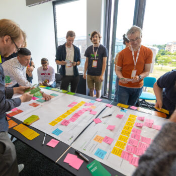 Dokumentation beim Barcamp: Teilnehmende stehen um einen Tisch, auf dem viele Post its kleben.