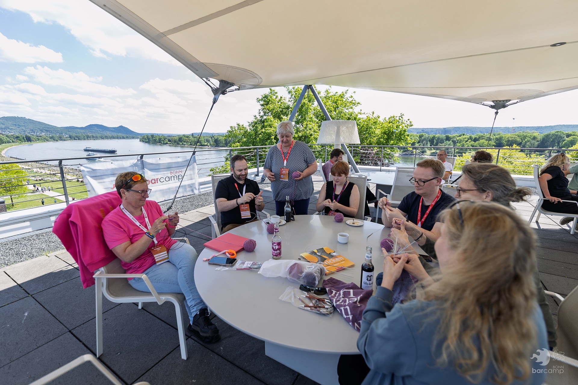 Gruppe von Menschen sitzt an rundem Tisch unter Sonnensegel auf Terrasse mit Fluss und Hügeln im Hintergrund